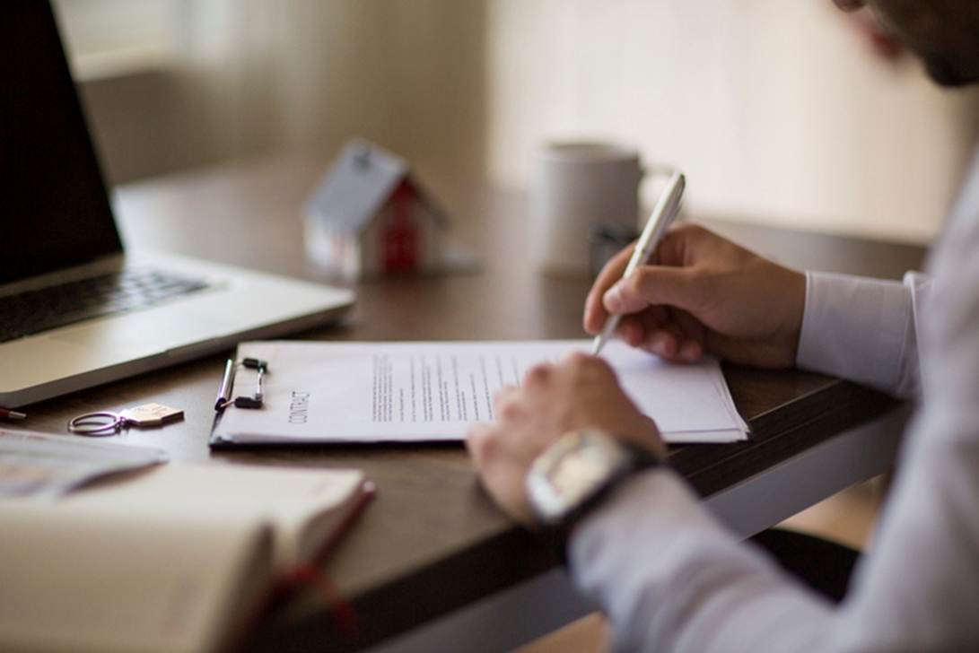 A person signs a contract with a pen, near a laptop, cup, and miniature house. The document is labeled "CONTRACT" in all caps on top.